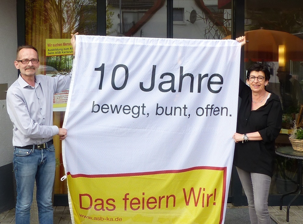 Bewegt, bunt, offen: Das ASB-Josefshaus feiert 10-Jähriges und eine Flagge macht das für alle sichtbar. Heimleiter Kurt Stahl hält sie zusammen mit Elke Weser-Hoffmann bereit. Die Pflegefachkraft, von Beginn an dabei, schätzt in dem Pflegezentrum in Ubstadt-Weiher „die Herzlichkeit“.